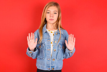 Serious blonde little kid girl wearing denim jacket over red background pulls palms towards camera,...