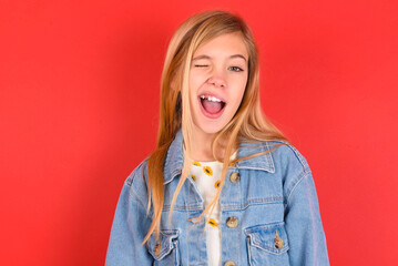 blonde little kid girl wearing denim jacket over red background winking looking at the camera with...