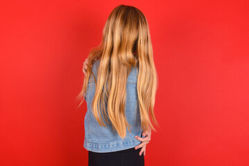 blonde little kid girl wearing denim jacket over red background hugging herself happy and positive from backwards. Self love and self care
