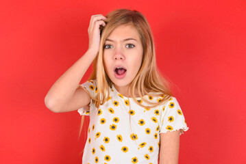 Embarrassed blonde little kid girl wearing daisy t-shirt red over background with shocked expression, expresses great amazement, Puzzled model poses indoor