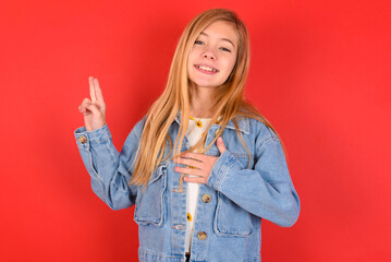 blonde little kid girl wearing denim jacket over red background smiling swearing with hand on chest and fingers up, making a loyalty promise oath.