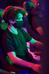 Focused young Caucasian man in mask and headset sitting in dark room and playing video game