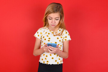 Excited blonde little kid girl wearing daisy t-shirt over red background winking and eye hold smart phone use read social network news