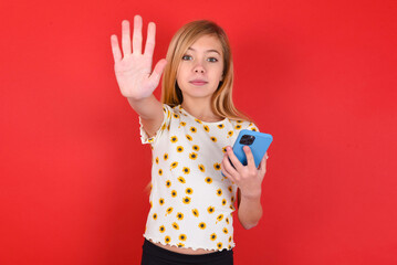 blonde little kid girl wearing daisy t-shirt over red background using and texting with smartphone...