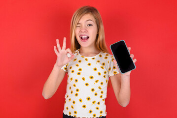 blonde little kid girl wearing daisy t-shirt over red background holding in hands cell showing...