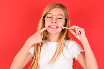 blonde little kid girl wearing denim jacket over red background holding an invisible aligner and pointing to her perfect straight teeth. Dental healthcare and confidence concept.