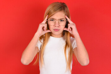 Serious concentrated blonde little kid girl wearing white t-shirt over red background keeps fingers on temples, tries to ease tension, gather with thoughts and remember important information for exam
