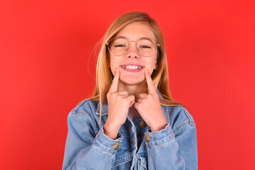 Happy blonde little kid girl wearing denim jacket over red background with toothy smile, keeps index fingers near mouth, fingers pointing and forcing cheerful smile