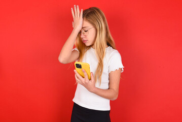 Upset depressed blonde little kid girl wearing white t-shirt over red background makes face palm as...