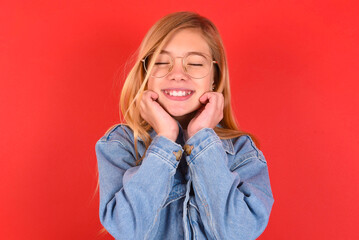 blonde little kid girl wearing denim jacket over red background grins joyfully, imagines something...