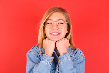 Satisfied blonde little kid girl wearing denim jacket over red background touches chin with both hands, smiles pleasantly, rejoices good day with lover