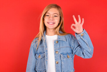 blonde little kid girl wearing denim jacket over red background hold hand arm okey symbol toothy...