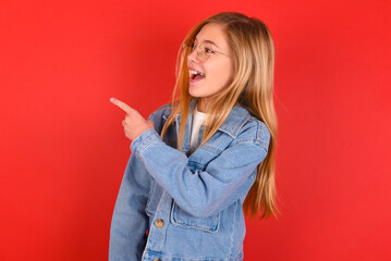 blonde little kid girl wearing denim jacket over red background glad cheery demonstrating copy...