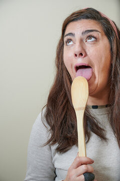 Young Adult Woman Gesticulates As She Tastes A New Recipe Using Wooden Cookware Spoon. She 
Is Licking The Spoon Looking Up.