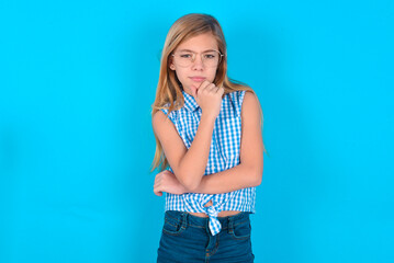 Thoughtful smiling little kid girl with glasses wearing plaid shirt over blue background keeps hand under chin, looks directly at camera, listens something with interest. Youth concept.