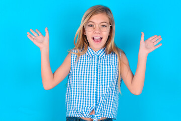 little kid girl with glasses wearing plaid shirt over blue background raising hands up, having eyes...