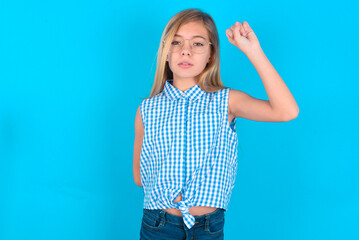 little kid girl with glasses wearing plaid shirt over blue background  feeling serious, strong and rebellious, raising fist up, protesting or fighting for revolution.