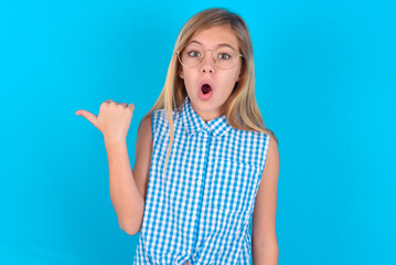 Shocked little kid girl with glasses wearing plaid shirt over blue background  points with thumb away, indicates something. Check this out. Advertisement concept.