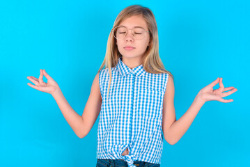 little kid girl with glasses wearing plaid shirt over blue background  doing yoga, keeping eyes...