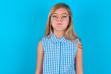 little kid girl with glasses wearing plaid shirt over blue background  puffing cheeks with funny face. Mouth inflated with air, crazy expression. - obrazy, fototapety, plakaty