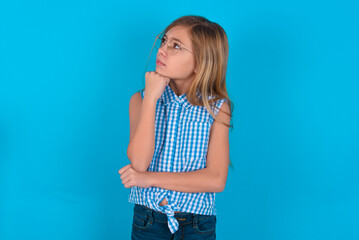 Portrait of thoughtful little kid girl with glasses wearing plaid shirt over blue back keeps hand under chin, looks away trying to remember something or listens something with interest. Youth concept.