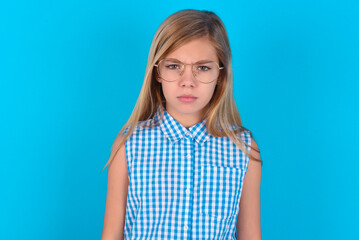 little kid girl with glasses wearing plaid shirt over blue background  frowning his eyebrows being displeased with something.