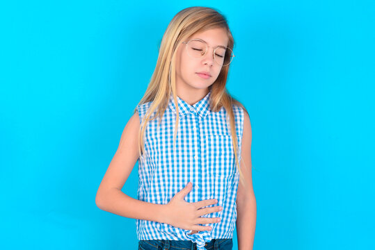 Satisfied Smiling Little Kid Girl With Glasses Wearing Plaid Shirt, Keeps Hands On Belly, Being In Good Mood After Eating Delicious Supper, Demonstrates She Is Full. Pleasant Feeling In Stomach.