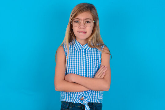 little kid girl with glasses wearing plaid shirt over blue background  bitting his mouth and looking worried and scared crossing arms, worry and doubt.