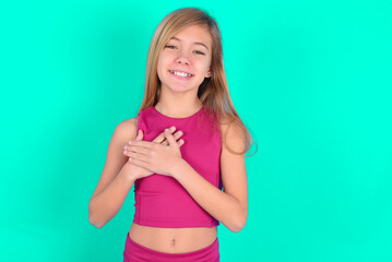 Honest blonde little kid girl wearing pink sport clothes over green background keeps hands on chest, touched by compliment or makes promise, looks at camera with great pleasure.