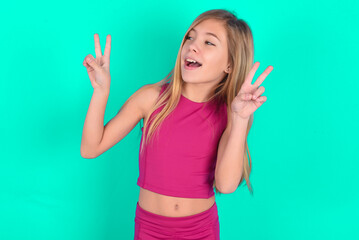 Isolated shot of cheerful blonde little kid girl wearing pink sport clothes over green background makes peace or victory sign with both hands, feels cool.