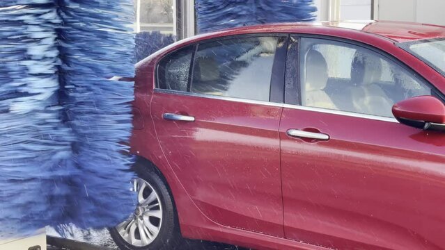 Sedan type red car going to the blue roller of the car wash, with foam on the bodywork, and the sun's rays breaking through the splashes of water