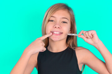 blonde little kid girl wearing black sport clothes over green background holding an invisible aligner and pointing to her perfect straight teeth. Dental healthcare and confidence concept.
