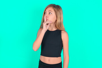 blonde little kid girl wearing black sport clothes over green background makes silence gesture,...