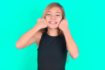 Fototapeta premium Happy blonde little kid girl wearing black sport clothes over green background keeps fists on cheeks smiles broadly and has positive expression being in good mood