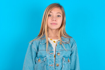 Stunned blonde little kid girl wearing denim jacket over blue background stares reacts on shocking...