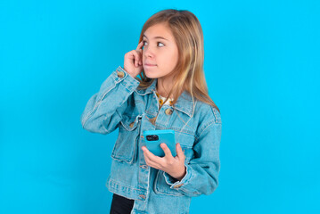 blonde little kid girl wearing denim jacket over blue background holding gadget while sticking out tongue