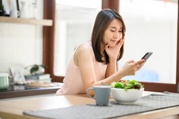 Obraz na płótnie Canvas Happy smiling Asian woman using smartphone mobile at home in kitchen with salad bowl on the table