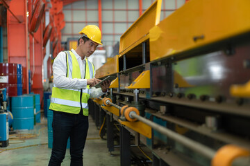 Asian male engineer wearing safety vest with helmet using tablet checking machine at factory...