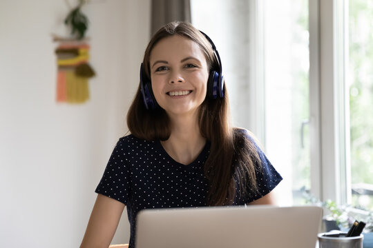 Woman in wireless headphones sit at desk with laptop staring at camera smile feels happy enjoy easy, comfort modern tech usage, e-learn, listen audio course to improve foreign language skill concept