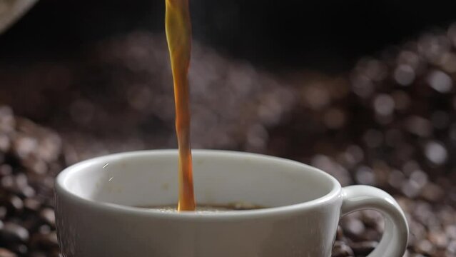 Pouring coffee with smoke on a cup and coffee beans piles on black background. Slow motion.