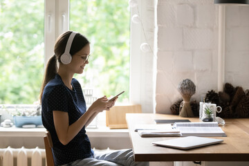 Woman in headphones studying sit at table with textbooks, take break holds smartphone share messages to friend, choose music through digital streaming services, use modern tech, hobby, leisure concept