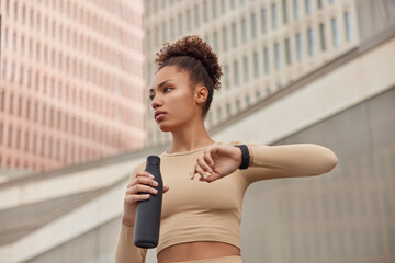 Waist up shot of healthy slim woman dressed in cropped top holds bottle of fresh water checks fitness results on smartwatch focused away stands against blurred background rests after workout