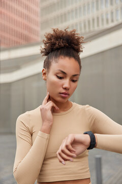Vertical shot of serious curly haired woman checks pulse on neck focused at smartwatch dressed in sportswear monitors heart rate from wrist poses outdoors. Sporty female uses workout tracker