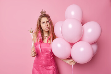 Obraz na płótnie Canvas Indoor shot of displeased indignant European woman wears crown and dress comes on costume party holds bunch of inflated balloons and magic wand discontent to hear bad news. Special occasion.