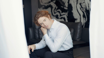 A man with a white dress shirt. Action. A man with red hair is sitting on the sofa and adjusting the collar of his clothes against a background of light black wallpaper and a bright white curtain in
