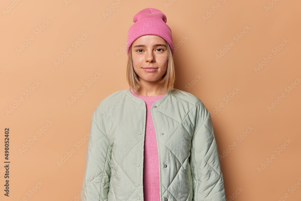 Wall mural Portrait of good looking blonde young woman looks directly at camera has confident expression wears hat and jacket going to have walk in park during leisure time isolated over beige background