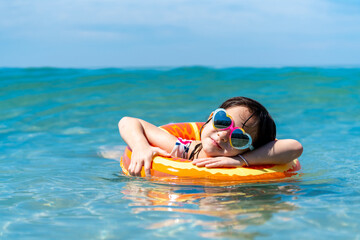  Little Asian child girl in swimwear with swim ring playing in sea water at tropical beach on summer sunny day. Happy children kid enjoy and fun outdoor activity lifestyle on beach holiday vacation
