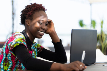 Cheerful young African woman following a tutorial from her favorite influencer blogger on her laptop, taking notes; global modern media consumption - obrazy, fototapety, plakaty