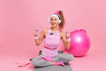 Obraz na płótnie Canvas Positive European woman dressed in sportsclothes stretches resistance band sits crossed legs on fitness mat uses sport equipment for exercising listens music via headphones poses against pink wall