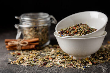 Herbal tea in a ceramic bowl and a glass jar on a black textural background. Detox and immunity tea. Herbal collection of chamomile, mint, lemon balm. rosehip and dried fruit pieces.